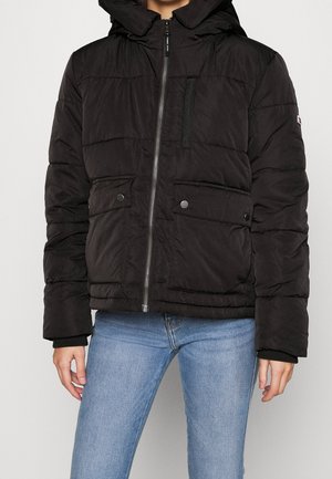 Persona che indossa una giacca a cappuccio nera imbottita con zip frontale e tasche, abbinata a jeans attillati di un azzurro chiaro.