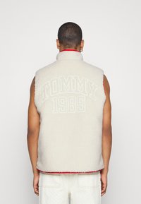 Gilet en polaire ivoire avec un col montant, des bordures rouges et une grande broderie "TOMMY 1985" au dos. Sans manches, texture douce et design sportif.