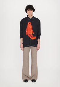 Black shirt with an orange cartoon dog graphic, structured design, long sleeves, and contrasting white stitching paired with beige flared trousers.