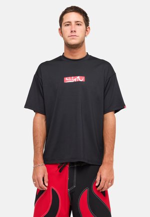 Mężczyzna ubrany w czarną oversize koszulkę z czerwono-białym logo oraz czerwono-czarne szorty w płomienie, stojący na jednolitym tle.