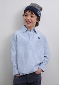 Ljusblå randig långärmad skjorta med knappkrage, prydd med en liten mörkblå skallelogotyp. Bärs med grå mössa och byxor.