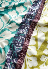 Tissu en coton avec un motif floral turquoise et blanc, accentué par des bordures à motifs violets et verts avec des détails intriqués.