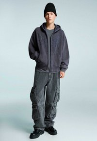 Mladostnik z črnim beaniejem, prevelikim faded črnim zip-up puloverjem z vezenim motivom, ohlapnimi cargo hlačami in črnimi debelimi čevlji, stoji.