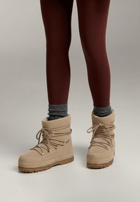 Beige mockaboots till ankeln med texturerad yta, rund tå, vadderad krage och snörning fram. Sulan är av gummi med mönstrat mönster för grepp.