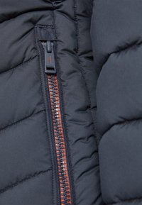 Detalj av en puffjacka som visar svart quiltat tyg, kontrasterande orange dragkedja med en plastdragflik och förstärkta sömmar längs kanterna.