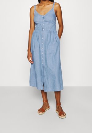 Frau trägt ein ärmelloses hellblaues Midi-Hemdkleid mit Taschen, kombiniert mit braunen Sandalen mit Knöchelriemen, steht vor weißem Hintergrund.