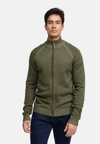 Sweater zippé vert olive avec un col haut, des poignets côtelés et un motif de tricot texturé. Présente un tirette de fermeture éclair marron avec un accent en cuir.