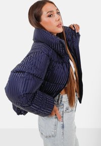 Giacca puffer blu navy con texture a coste verticali, colletto alto, chiusura con zip e maniche oversize, abbinata a jeans in denim chiaro.