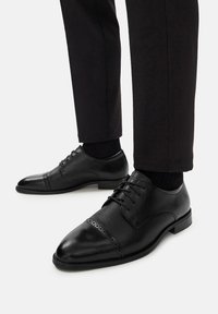 Czarne skórzane buty do garnituru z perforowanym wzorem na przodzie, zaokrąglonym noskiem i sznurowadłami. Noszone z ciemnymi spodniami i czarnymi skarpetkami.