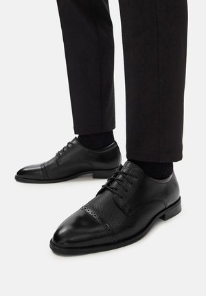 Melnas ādas kleitu kurpes ar perforētu dizainu priekšpusē, noapaļota purngala un auklām. Valkātas ar tumšiem biksēm un melnām zeķēm.