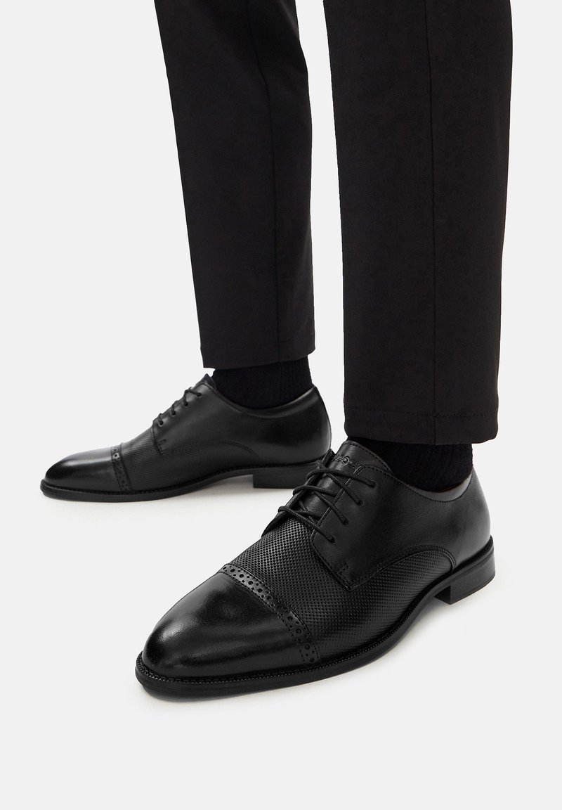 Czarne skórzane buty do garnituru z perforowanym wzorem na przodzie, zaokrąglonym noskiem i sznurowadłami. Noszone z ciemnymi spodniami i czarnymi skarpetkami.