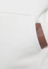 Sudadera blanca con bolsillo canguro, tela texturizada y puños acanalados. Una mano de piel morena está ubicada dentro del bolsillo.