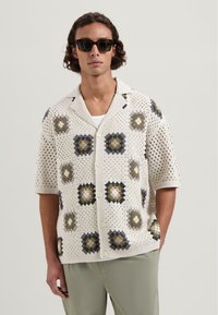Korte mouwen haakshirt in crème, met geometrische patronen in donkerblauw, mosterd en grijs. Verschlossen voorkant en een relaxte pasvorm.