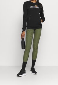 Czarna bluza z logo, legginsy w zielone paski i czarne sneakersy. Model trzyma czarną butelkę na wodę. Prosty, sportowy styl.