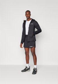 Black athletic jacket with hood and logo, paired with black shorts. Model wears white t-shirt and dark sneakers with light accents.