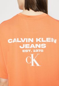 Camiseta de algodón naranja con texto en blanco: "CALVIN KLEIN JEANS EST. 1978" y el logo "ck". Diseño simple y holgado con cuello redondo.