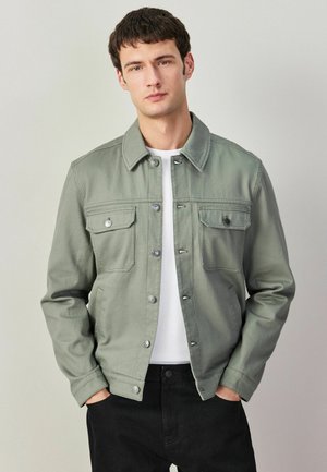 Młody mężczyzna w jasnoszaro-zielonej kurtce na guziki, noszący białą koszulę i czarne spodnie, stojący z rękami w kieszeniach na jednolitym tle.