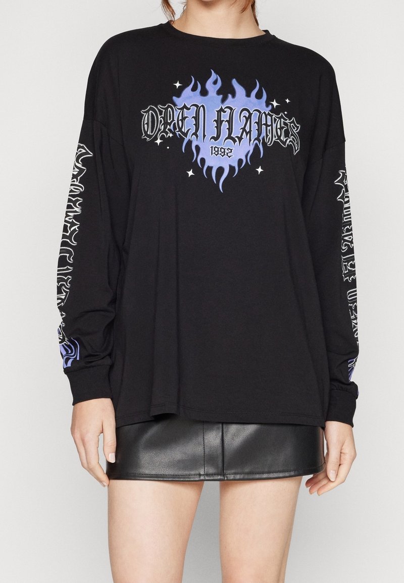 Svart långärmad t-shirt med blå eld grafik, texten "OPEN FLAMES 1992" och vita mönster på ärmarna; kombinerad med en svart läderkjol.