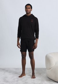 Μαύρο φούτερ με κόκκινη καρδιά λογότυπο και κείμενο, συνδυασμένο με μαύρο σορτς που ταιριάζουν. Και τα δύο ρούχα έχουν άνετη εφαρμογή και μαλακή υφή.