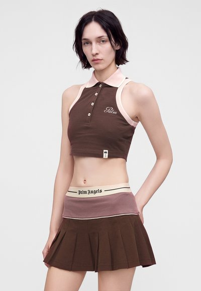 Γυναίκα που φοράει καφέ cropped polo τοπ με ροζ περιθώριο και καφέ πλισέ φούστα με φαρδιά μέση που γράφει "Palm Angels."