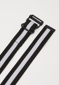 Svart og hvit stripete belte laget av vevd nylon, med en robust svart plastspenne og justerbar lengde.
