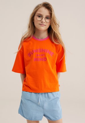 Plavuša s okruglim naočalama, u narančastoj majici s natpisom „SUNSHINE ON MY MIND“ i svijetloplavim hlačicama, stoji na bež pozadini.