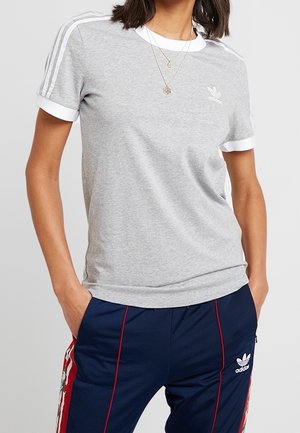 Femme portant un t-shirt gris Adidas avec des bandes blanches sur les manches et un pantalon Adidas bleu marine avec un liseré rouge et des bandes florales sur les côtés.