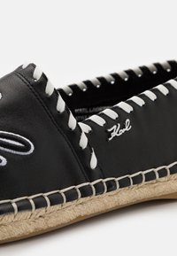 Espadrille en cuir noir avec coutures blanches, semelle en jute tressée et logo « Karl » argenté sur le côté.