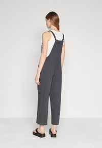 Gray, textured overalls with adjustable straps, straight leg design, and side pockets, worn over a white top and paired with black sandals.