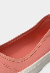 Gros plan d'une chaussure en toile couleur corail rose, sans lacets, avec une semelle en caoutchouc blanche, montrant l'intérieur de la chaussure et les détails de la couture.