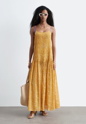 Femme portant une robe longue jaune à fleurs avec des bretelles violettes, des lunettes de soleil blanches, des sandales, des bracelets et tenant un sac cabas tissé beige.