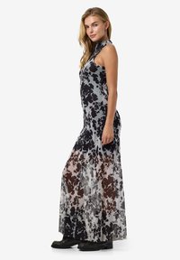 Rochie maxi cu un model abstract gri și negru, design fără mâneci și material transparent. Lungime până la podea, cu un corset ajustat. Încălțăminte neagră.