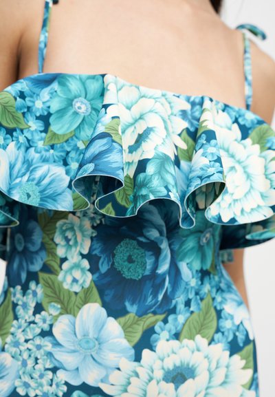 Robe bleue fleurie à épaules dénudées avec un col volanté et fines bretelles, montrant le haut de la poitrine et une partie du torse.