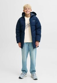 Plavi dečko se smije, nosi tamnoplavu zimskojaknu preko krem majice Jack & Jones, svijetloplave traperice i sive tenisice, stoji na bijeloj pozadini.