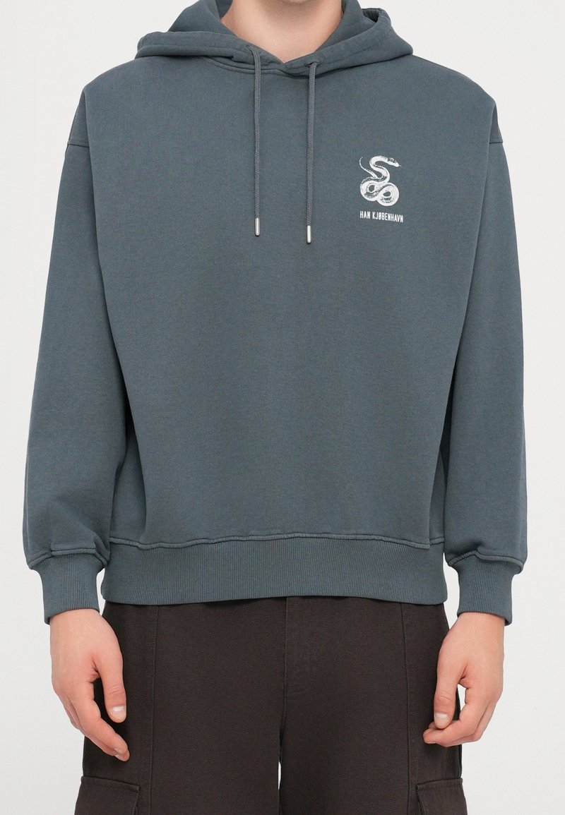Personne portant un sweat à capuche gris avec un logo de serpent blanc et le texte « HAN KJØBENHAVN » sur le haut à gauche de la poitrine.