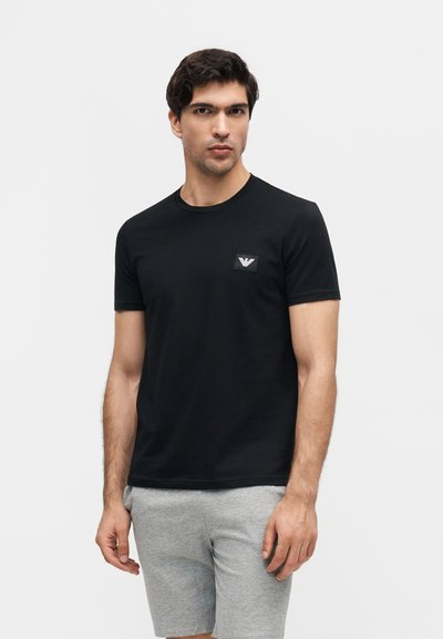 Άνδρας που φοράει μαύρη κοντομάνικη μπλούζα με μικρό λογότυπο στο στήθος και ανοιχτό γκρι σορτς, στέκεται μπροστά σε απλό λευκό φόντο.