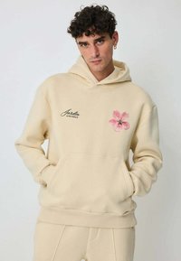 Sweat à capuche beige en tissu doux, doté d'une poche avant, d'une capuche avec cordon de serrage et d'un motif floral rose sur la poitrine gauche avec le texte "Jardin de Provence".