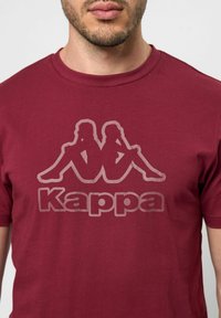 Burgundi színű pamut póló, amelyen egy nagy, elhalványult Kappa logó és árnyékos grafika található az elején. Normál, kerek nyakú és rövid ujjú.