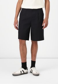 Black pleated shorts, knee-length, made of lightweight fabric. Paired with white sneakers featuring black stripes and black socks.