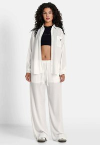 Witte, gestructureerde overhemd met knopen over een zwart cropped top, gecombineerd met losse witte broek. Bevat een zakdetail en een koordjes taille.