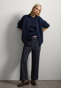 Donkerblauwe gebreide cardigan met grote knopen, gecombineerd met een donkere trui, gestructureerde grijze broek en bruine veterschoenen.
