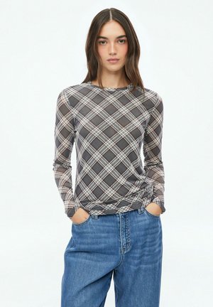 Jeune femme aux longs cheveux bruns portant une chemise grise à carreaux à manches longues et un jean bleu ample, debout les mains dans les poches.