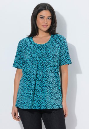 Kvinde med glat sort hår iført en blå kortærmet top med et lille sort blomsterprint og sorte bukser mod en hvid baggrund.