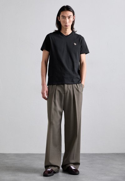 T-shirt en coton noir à col en V avec un petit logo, associé à un pantalon beige ample avec des plis et des chaussures marron foncé avec des lacets blancs.