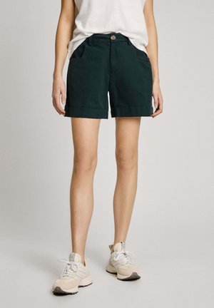 Dunkelgrüne Baumwollshorts mit flachem Bund, umgeschlagenem Saum und Seitentaschen, kombiniert mit hellen Turnschuhen.