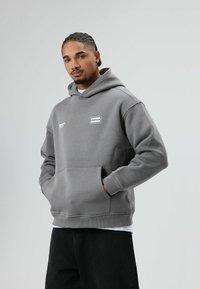 Grijze capuchontrui met een kangoeroezak en ribgebreide manchetten. Voorzien van een witte logopatch op de borst. Draagt over een wit T-shirt.