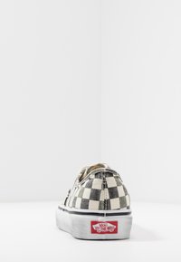 Chaussure slip-on en toile à carreaux noirs et blancs avec une semelle en caoutchouc et une étiquette avec logo rouge à l'arrière. Présente une pointe arrondie et des accents élastiques sur les côtés.