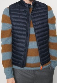 Mørkeblå quiltet vest med høj krave, lynlås og orange lynlåstræk, båret over en stribet sweater i blå og brune nuancer.