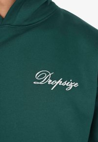 Dunkelgrüner Kapuzenpullover mit weißer, geschwungener "Dropsize"-Stickerei auf der linken Brust.