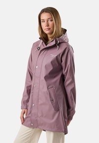 Kvinde iført en mosefarvet vandtæt hættetrøje med knaplukning på lommerne og cremefarvede bukser, stående mod en hvid baggrund.